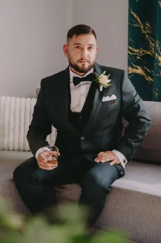 Pan młody ze szklanką whisky siedzący na kanapie w butonierce z białą różą