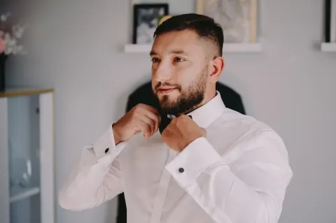 Pan młody poprawia muchę przed lustrem w białej koszuli ze spinkami