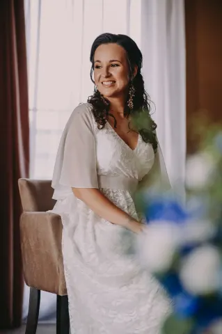 Roześmiana panna młoda w zwiewnej sukni przy oknie, w tle bukiet kwiatów
