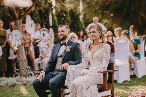 Para młoda siedzi na drewnianych krzesłach podczas ceremonii w ogrodzie