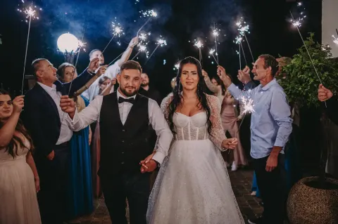 Para młoda wychodzi w szpalerze gości z zimnymi ogniami po zmroku